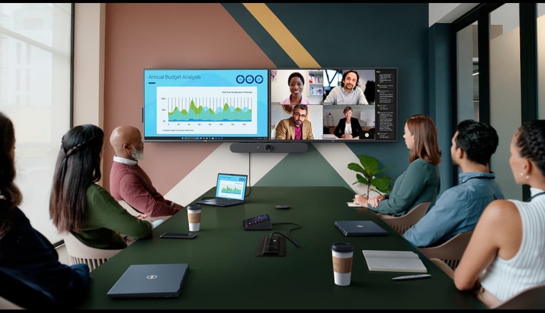 Professionals in a conference room viewing a budget chart and video call on dual screens.