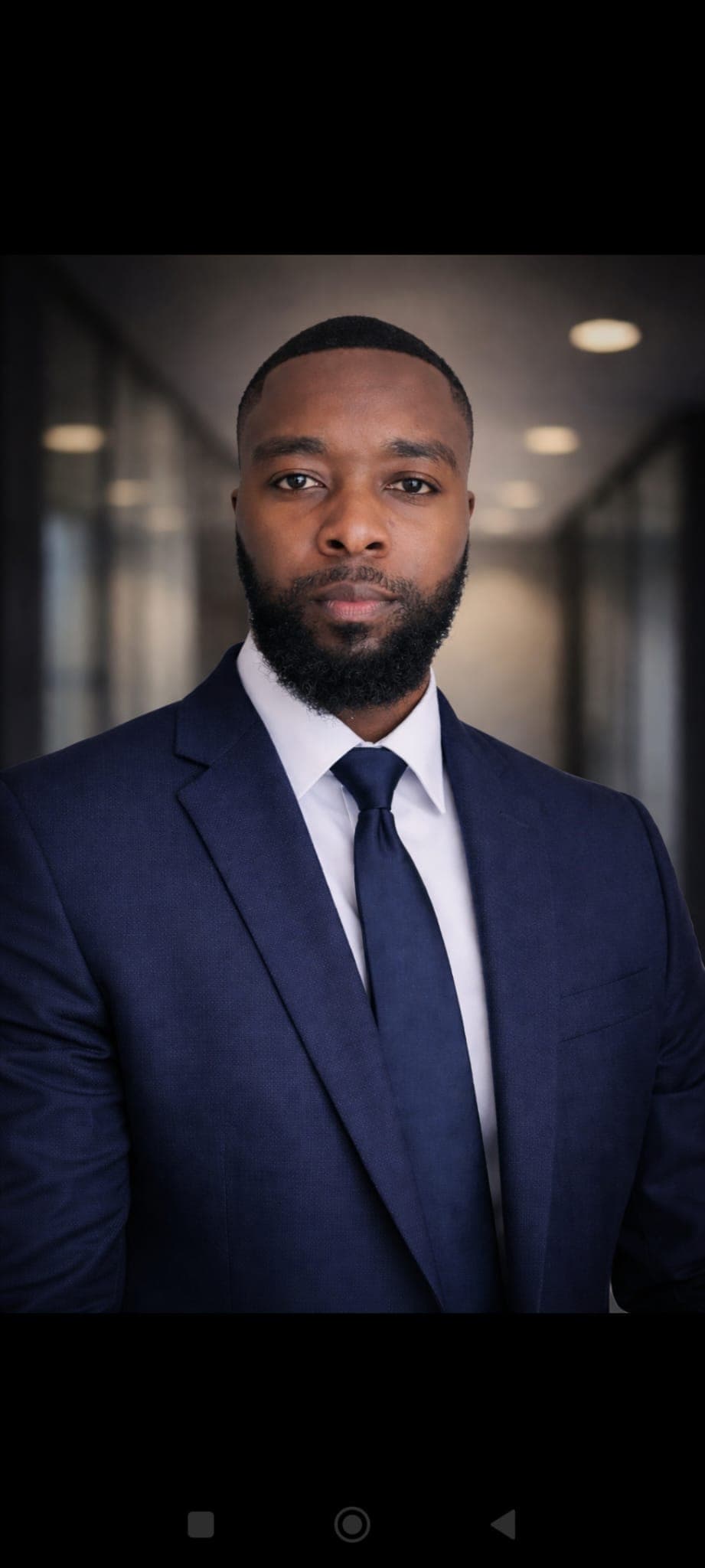 Bearded Black man in a navy blue suit and tie against a blurred office background.